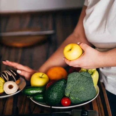 Woman grabs a yellow apple from a plate of healthy foods while pushing away a plate of desserts
