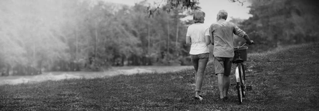 Older couples in black and white walks through park with bicycle