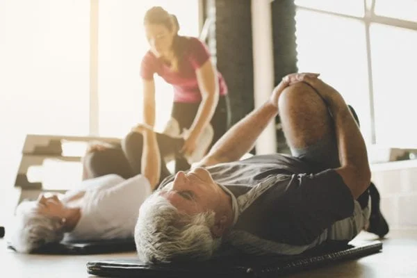 Older couples stretches on mats while therapist assists the woman